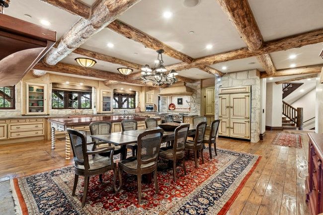 Spacious kitchen and dining area with exposed wood ceiling beams, wood-finish flooring, custom cabinetry, stone accent walls, and an integrated paneled refrigerator