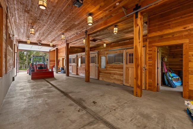 Interior barn area featuring wood-paneled walls and ceiling, concrete flooring with a drainage system, recessed lighting, and multiple enclosed stalls with sliding wood doors and metal grilles
