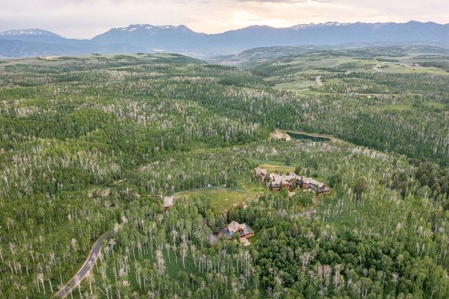 Expansive forested landscape featuring multiple structures, a winding paved road, and a calm body of water