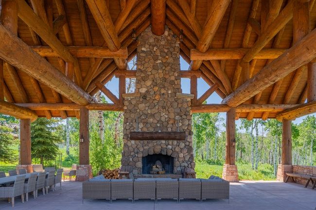 Expansive covered outdoor living area featuring a prominent stone fireplace, vaulted wood-beam ceiling, and stone-clad columns