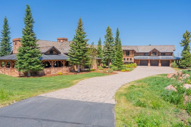 Expansive log-style residence featuring a multi-car garage, stone masonry accents, a paved driveway, and mature evergreen trees