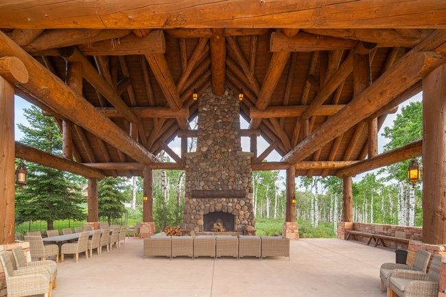 Expansive covered patio featuring a stone fireplace, exposed timber beam ceiling, integrated stone benches, and concrete flooring
