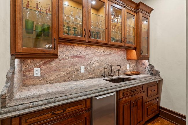 Built-in wet bar featuring custom wood cabinetry with glass-front uppers, under-cabinet lighting, a stone countertop, and a matching stone backsplash