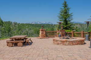 Expansive paver patio featuring a built-in stone fire pit, wood railing, and a mature evergreen tree
