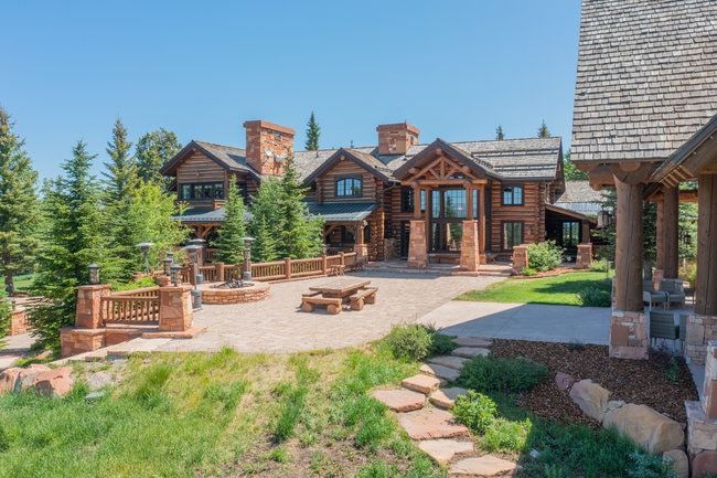 Expansive log home with stone chimneys, featuring a flagstone patio, built-in fire pit, and mature evergreen landscaping