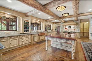 Rustic kitchen featuring exposed wood beams, light-toned custom cabinetry, wood-finish flooring, a central island with a polished countertop, and a built-in copper farmhouse sink