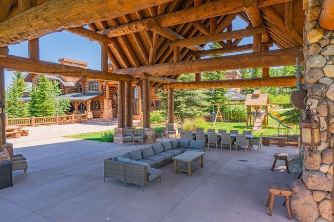 Expansive covered patio featuring a robust log beam ceiling, stone pillars, concrete flooring, and an adjacent log home with a wood railing and mature trees