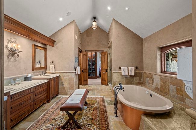 Spacious bathroom featuring a copper-tone soaking tub, stone tile flooring, a dual vanity with light-colored countertops, wood-finish cabinetry, and a vaulted ceiling with recessed lighting