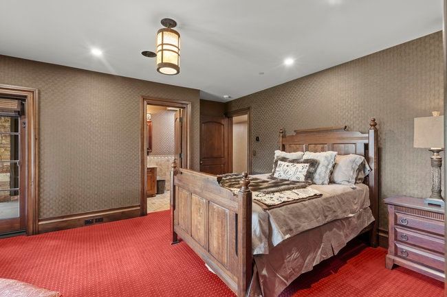 Recessed lighting and a central pendant fixture illuminate a room featuring patterned wallpaper, red textured carpeting, and detailed wood trim
