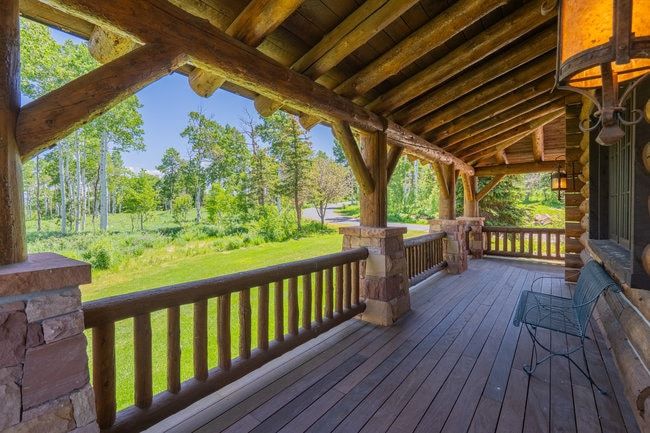 Expansive covered porch featuring a log beam ceiling, wood plank flooring, stone-clad pillars, log railings, and a log exterior wall