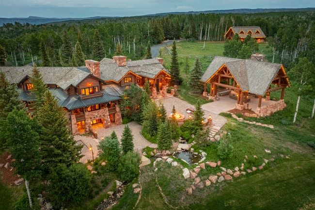 Expansive stone and log construction residence featuring multiple gabled roofs, an outdoor covered pavilion with exposed timber framing, a stone fire pit, and a naturalistic water feature