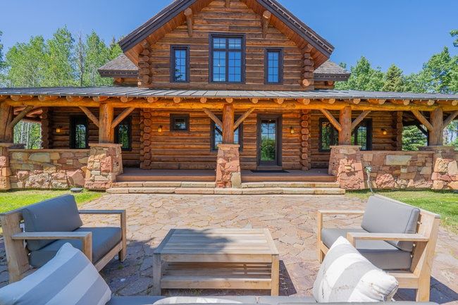 Exterior log home featuring a covered front porch, stone accents, multiple windows with dark frames, and a textured stone patio