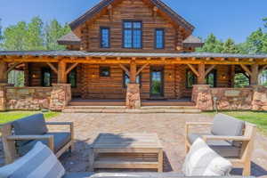 Exterior log home featuring a covered front porch, stone accents, multiple windows with dark frames, and a textured stone patio