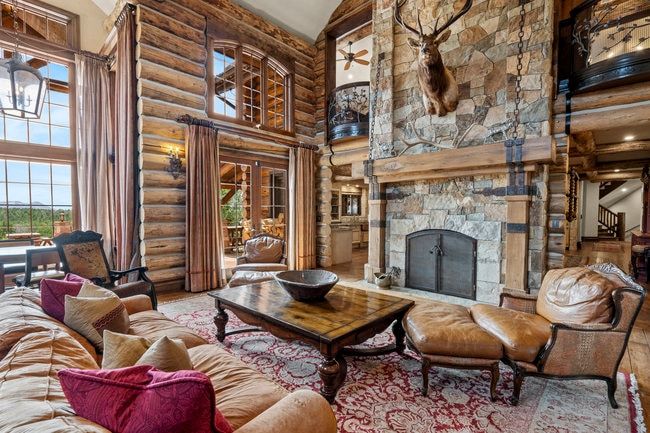 Great room featuring log wall construction, a stone fireplace with a substantial wood mantel, a double-height window wall with transom windows, and a balcony with an ornate railing