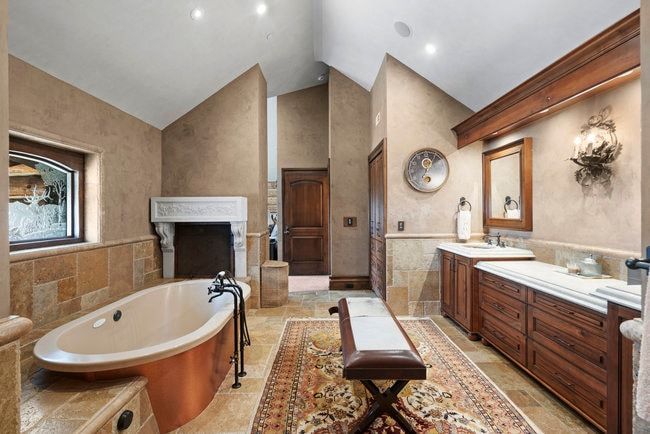 Freestanding soaking tub with floor-mounted faucet hardware, stone tile flooring and wainscoting, a decorative fireplace surround, and a dual vanity with undermount sinks and wood cabinetry