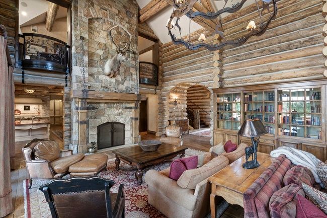 Grand living space featuring an impressive stone fireplace with a wood mantle, log wall construction, a built-in bookcase with glass doors, and a custom wrought iron chandelier
