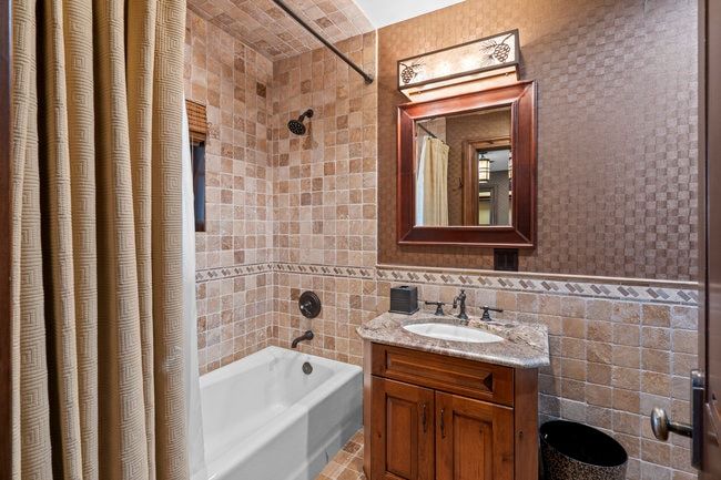Bathroom with tile surround tub/shower combination, decorative tile wainscoting, a vanity with stone countertop, and a framed mirror with an overhead light fixture