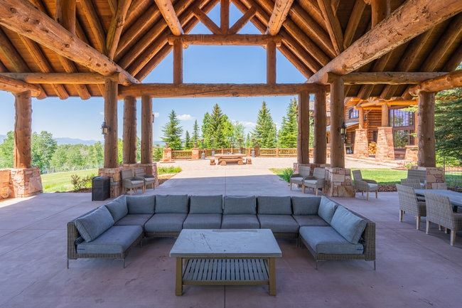 Spacious covered patio with exposed log beam construction, stone column bases, integrated wall sconces, and a concrete surface