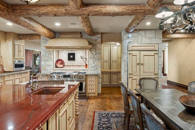 Gourmet kitchen featuring a large wood-finish island with an integrated sink, custom cabinetry with ornate hardware, professional-grade stainless steel range with custom hood, stone accent walls, and exposed wood beams