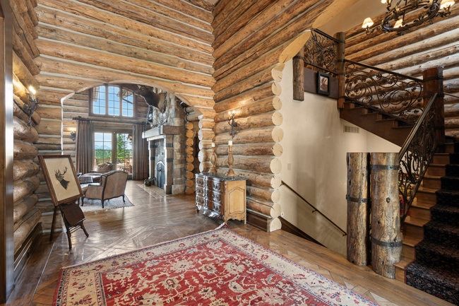 Rustic log cabin interior featuring exposed log walls, wood-finish flooring, a grand staircase with ornate wrought iron railings, and a stone fireplace