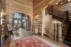 Rustic log cabin interior featuring exposed log walls, wood-finish flooring, a grand staircase with ornate wrought iron railings, and a stone fireplace