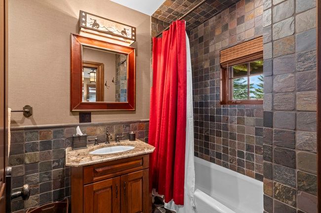 Bathroom featuring stone-finish wall tiling, a wood-frame vanity with a granite-finish countertop, and a built-in bathtub with an overhead shower