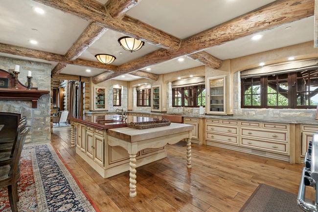 Expansive kitchen featuring exposed wood beam ceilings, wood-finish flooring, a substantial center island with butcher block and stone countertops, custom cabinetry with glass-front display units, and stone wall accents