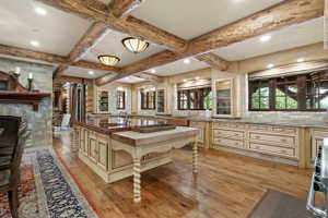 Expansive kitchen featuring exposed wood beam ceilings, wood-finish flooring, a substantial center island with butcher block and stone countertops, custom cabinetry with glass-front display units, and stone wall accents