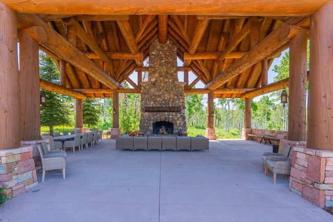 Expansive covered patio featuring a stone fireplace, exposed timber beams, a vaulted wood ceiling, concrete flooring, and stone pillar bases