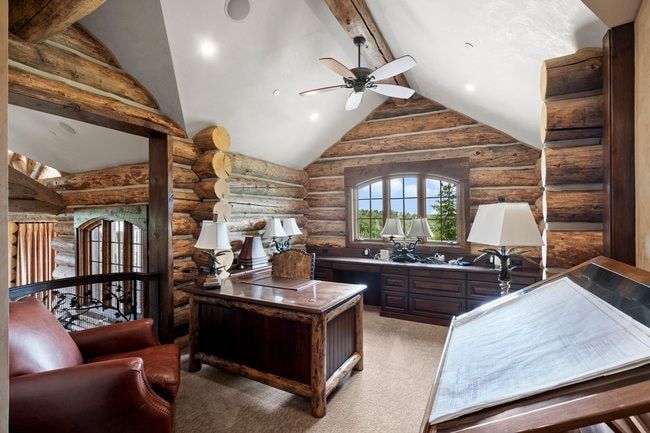 Log cabin-style office featuring exposed log walls, a built-in wooden desk, arched window with outdoor scenery, and a ceiling fan