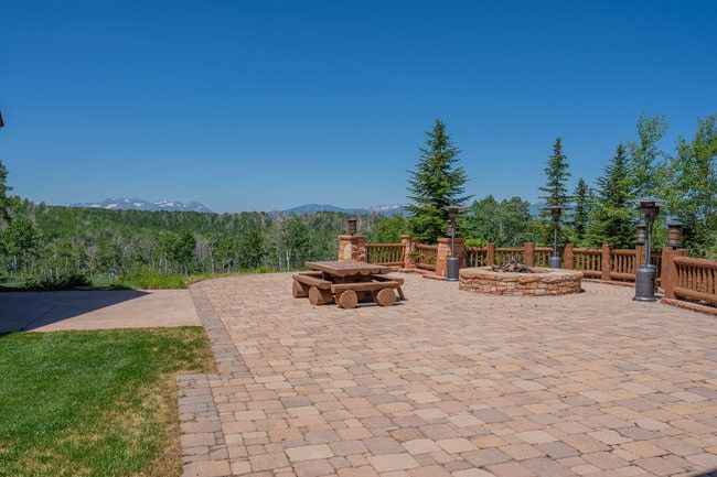 Expansive paver patio featuring a built-in stone fire pit, log railing, and mature evergreen trees
