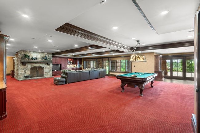 Spacious recreation room featuring a stone fireplace with hearth, red patterned carpeting, recessed lighting, and dark wood ceiling beams
