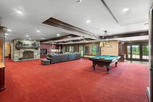 Spacious recreation room featuring a stone fireplace with hearth, red patterned carpeting, recessed lighting, and dark wood ceiling beams