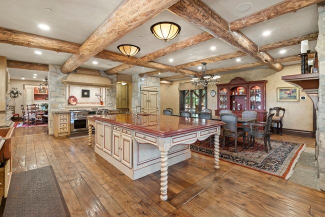 Expansive kitchen and dining area featuring wood-finish flooring, exposed wood beam ceiling, central island with storage and integrated sink, stainless steel range, and stone accents