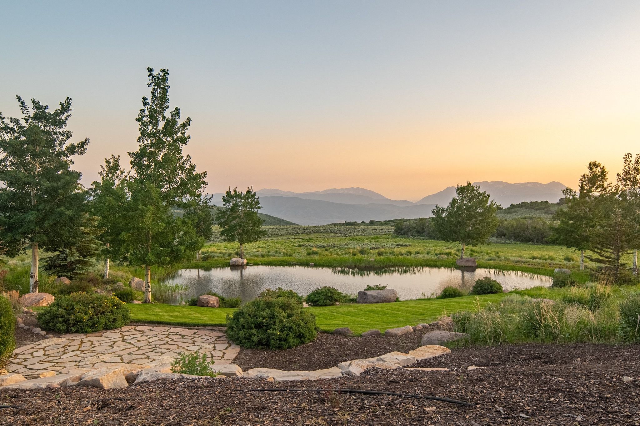 Expansive pond with surrounding natural landscape, mature trees, a stone patio, and distant mountain ranges