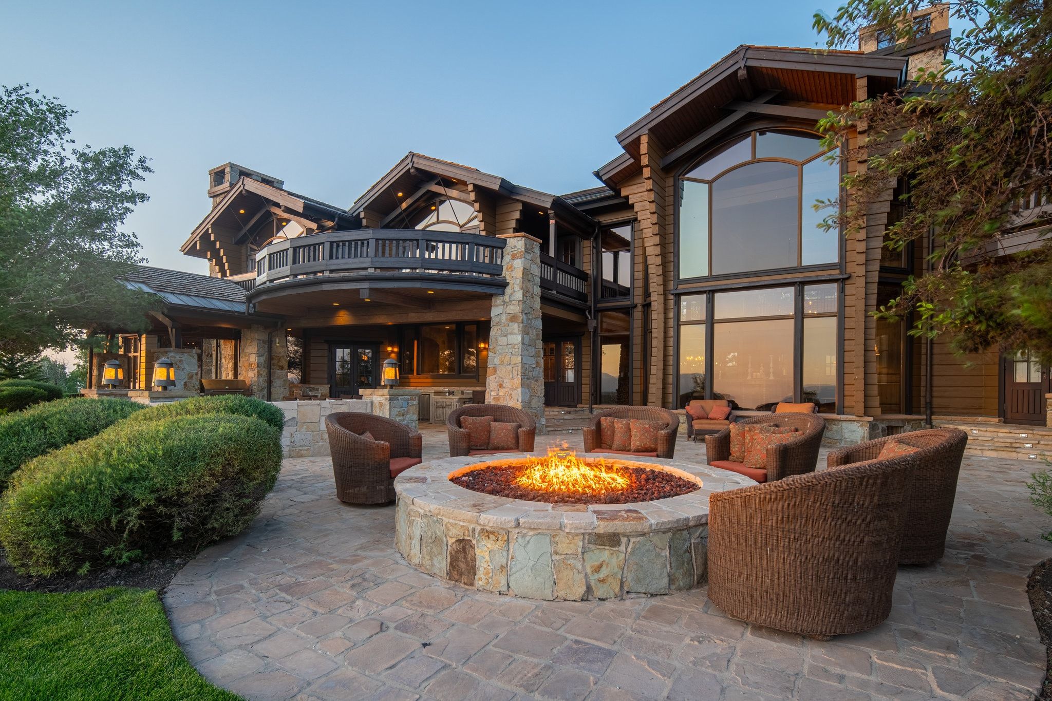 Expansive stone patio with a built-in stone fire pit, a curved upper-level balcony, large picture windows, and a log cabin style exterior