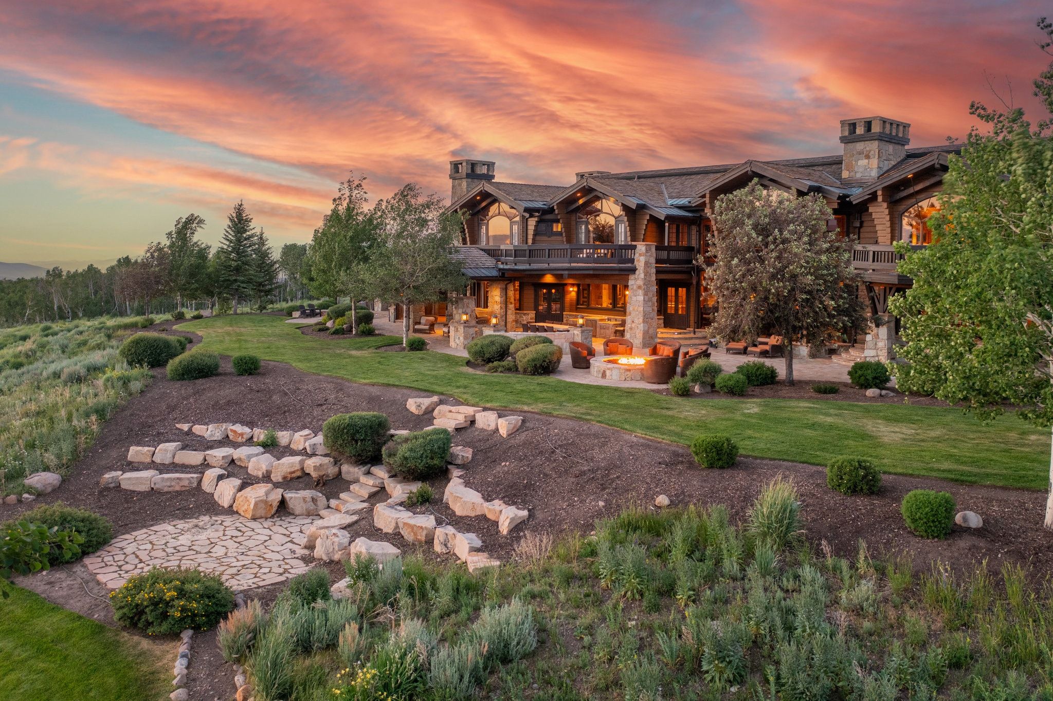 Expansive multi-level property featuring a stone and timber exterior, multiple balconies, a spacious stone patio, an outdoor fire pit area, and landscaped grounds with mature trees and stone hardscaping
