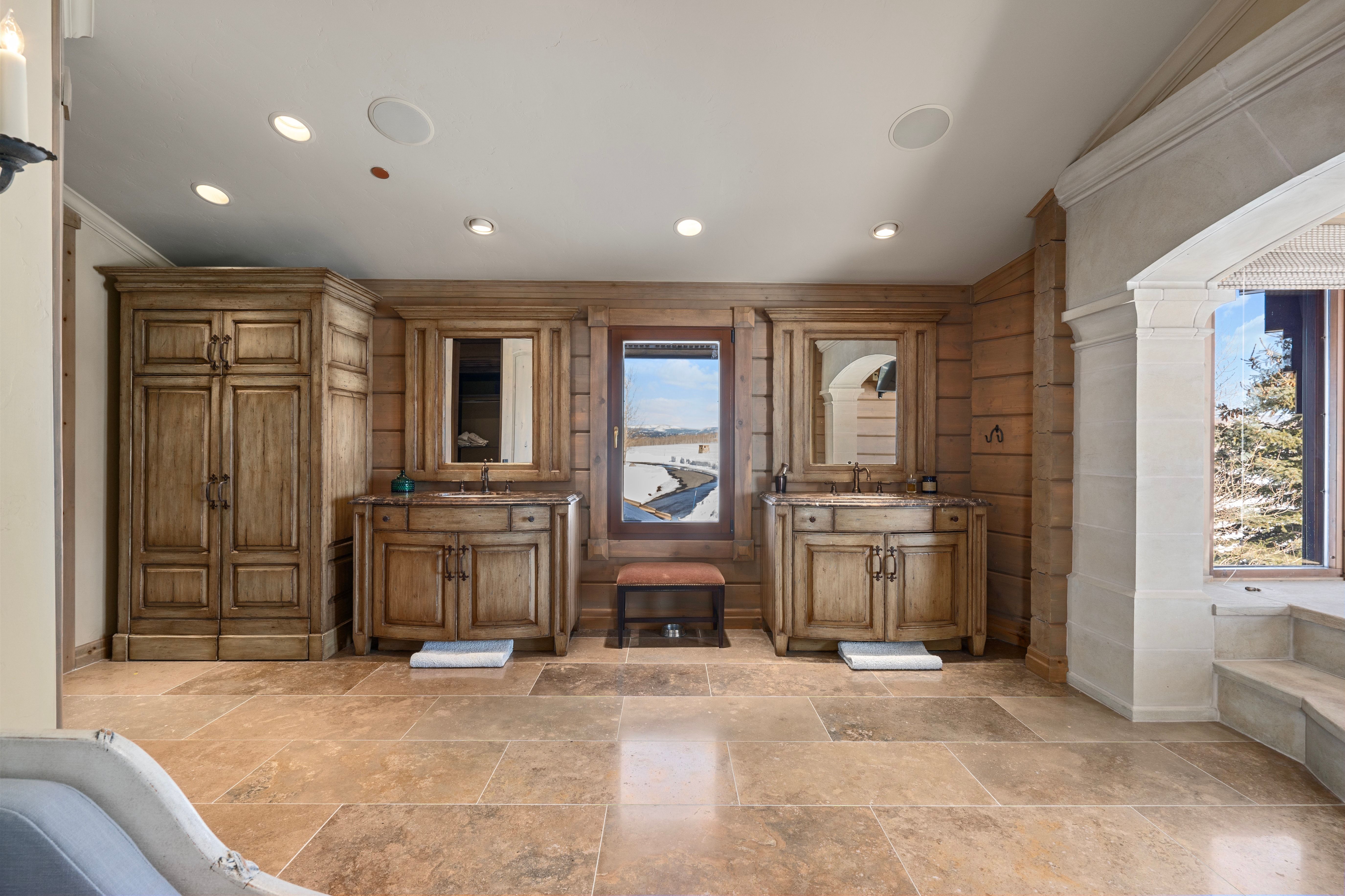 Spacious room featuring extensive stone tile flooring, custom wood cabinetry, dual vanity areas with integrated sinks, and ample natural light from multiple windows