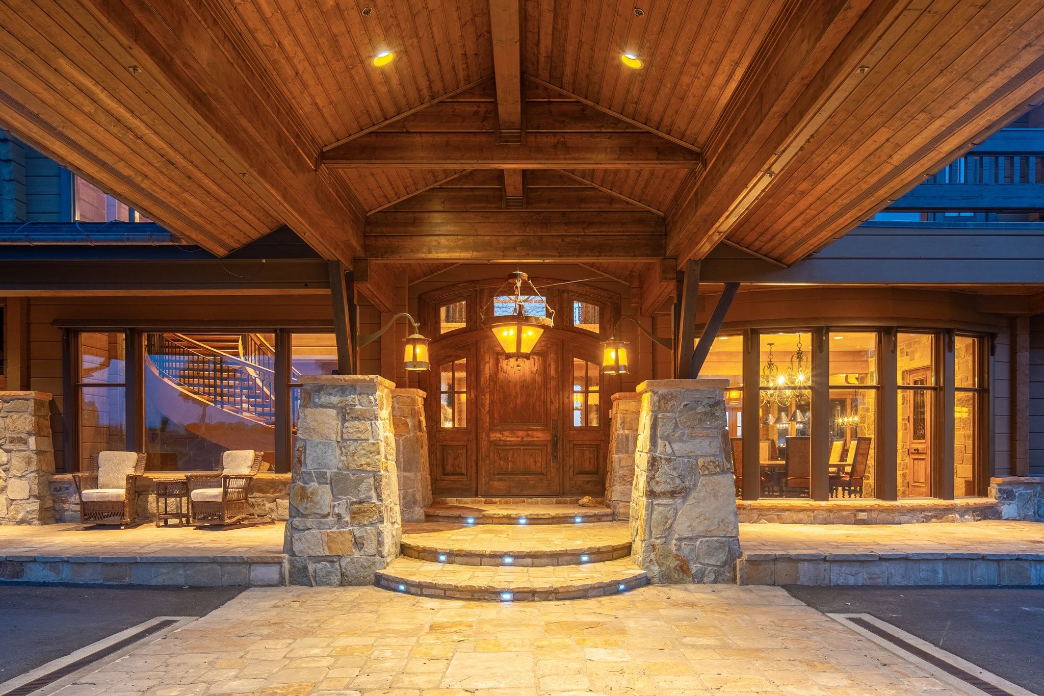 Stone and timber entry featuring a covered porte-cochère, decorative double doors, integrated step lighting, a stone-faced foundation, and an outdoor patio