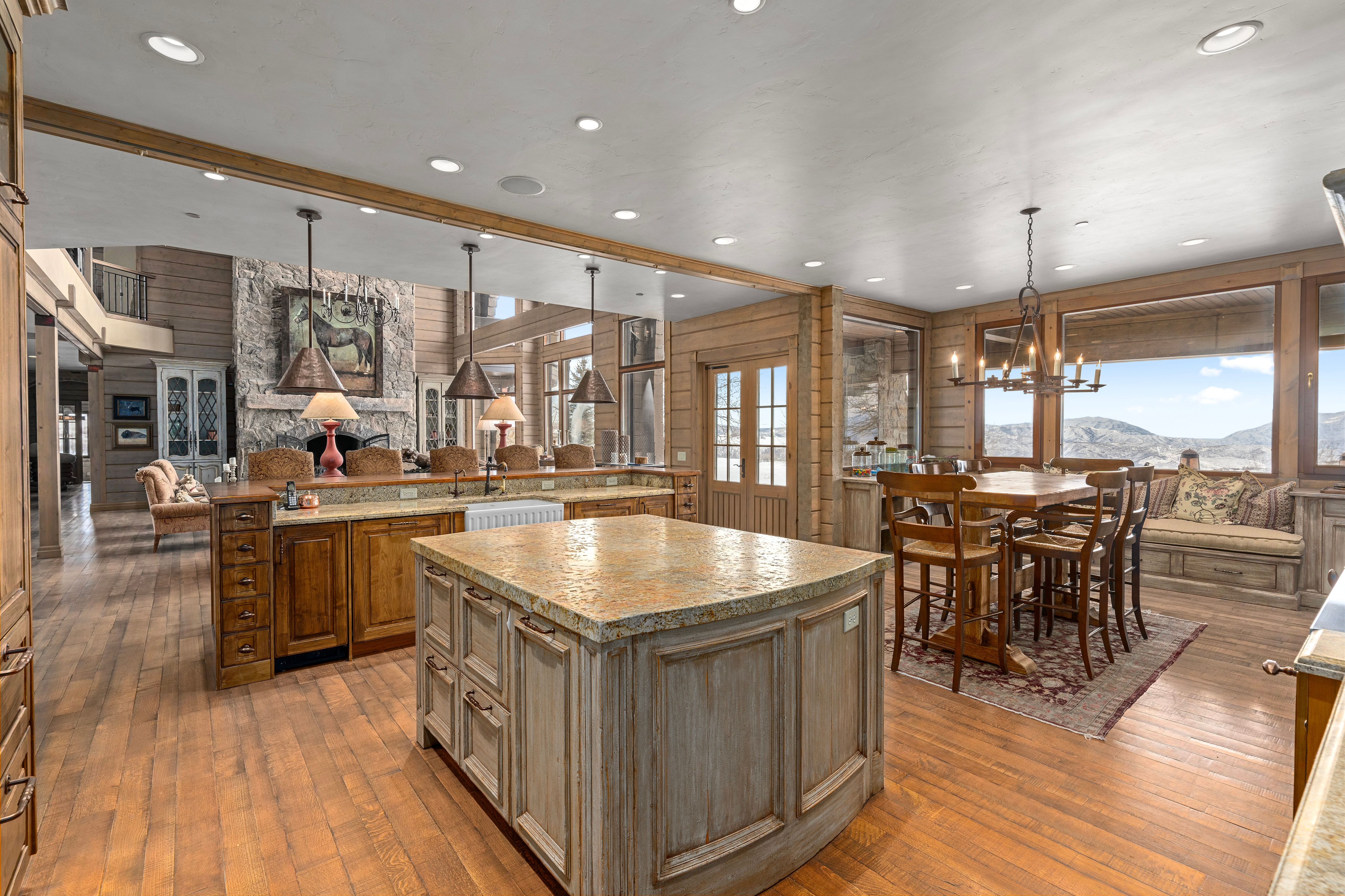 Expansive kitchen and dining area featuring wood-finish flooring, two large kitchen islands with stone countertops, a farmhouse sink, and panoramic windows showcasing mountain scenery