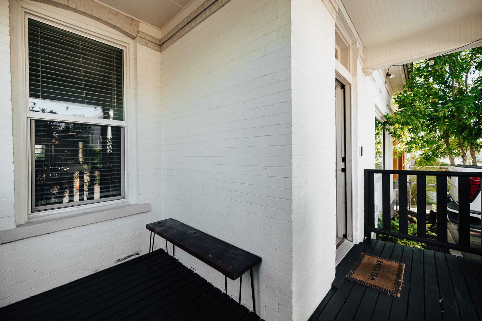 Porch entry featuring a painted brick exterior, white framed window with blinds, painted wood decking, and a black wood railing