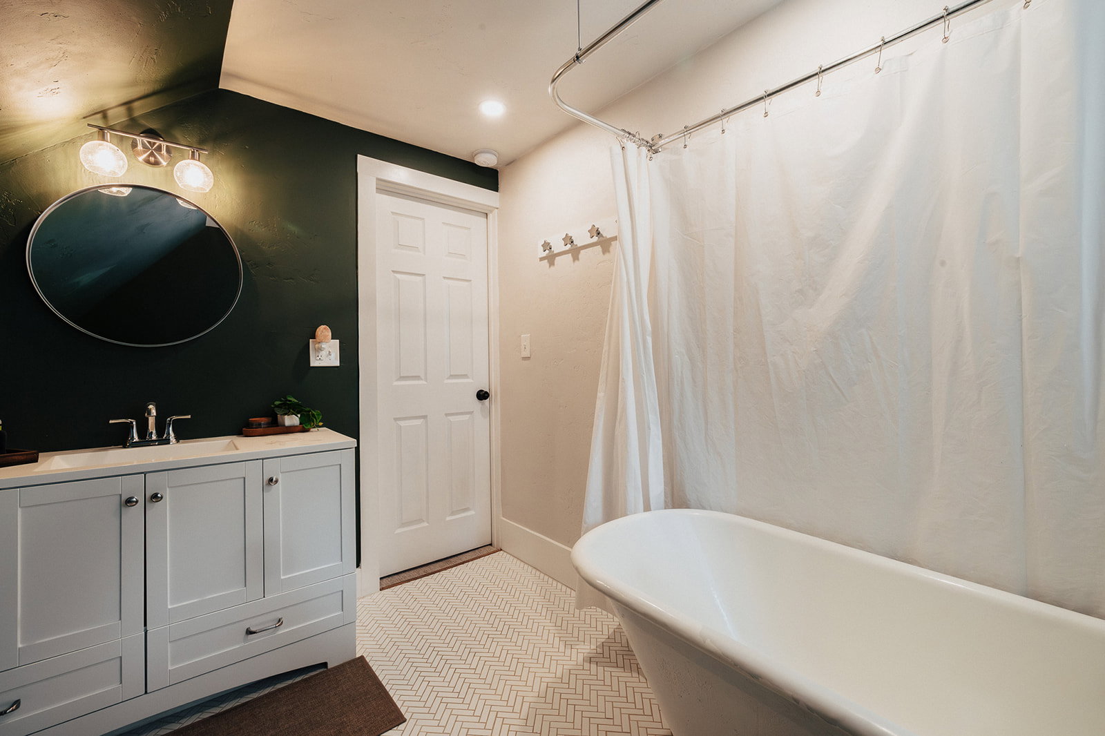Bathroom featuring a freestanding bathtub, white herringbone tile flooring, a double vanity with a light gray cabinet and white countertop, a circular mirror, and a dark green accent wall