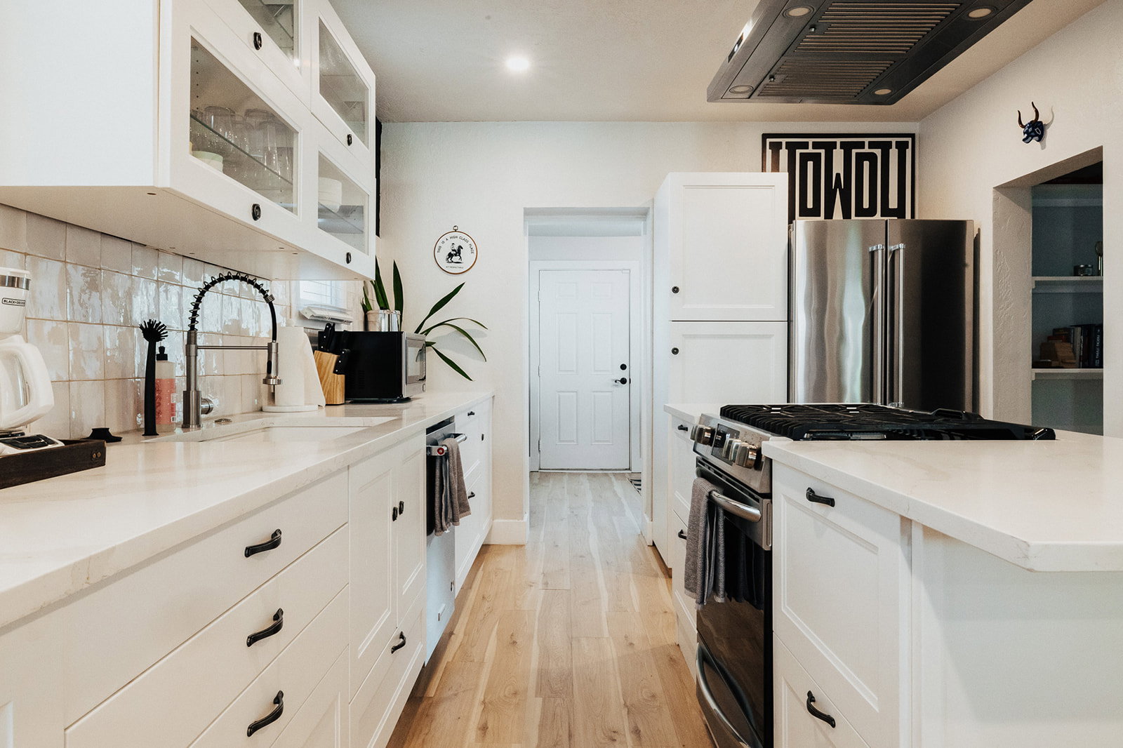 Modern kitchen featuring light wood-finish flooring, white cabinetry with dark hardware, a white tile backsplash, a stainless steel refrigerator, and a built-in range