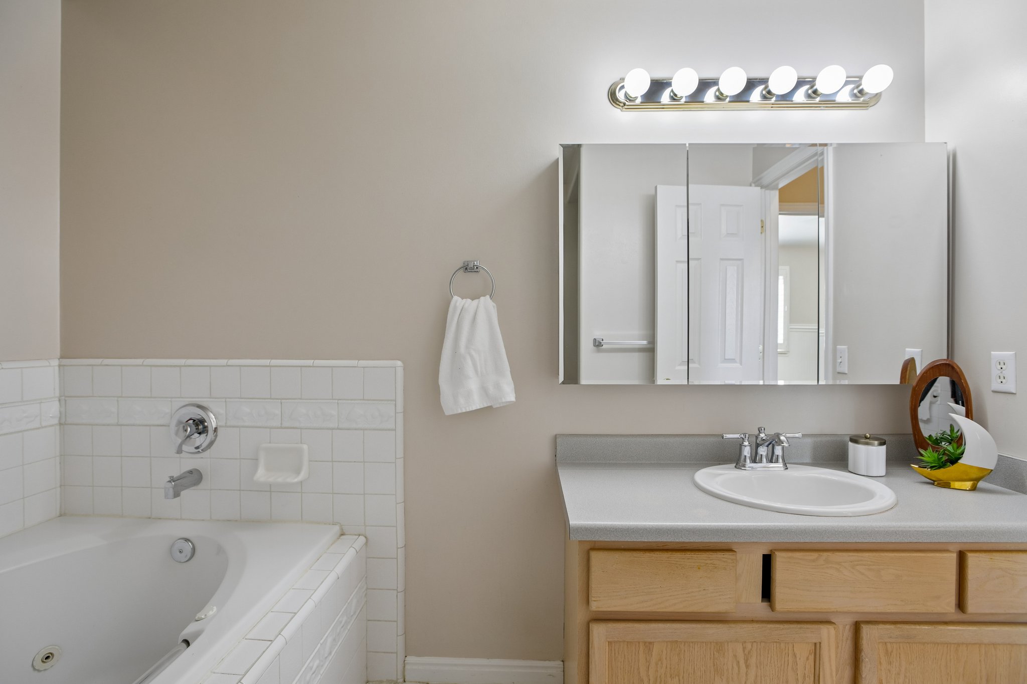 Bathroom featuring a white tiled soaking tub, a vanity with a laminate countertop, an integrated sink, natural wood-finish cabinetry, and a mirrored medicine cabinet with overhead lighting