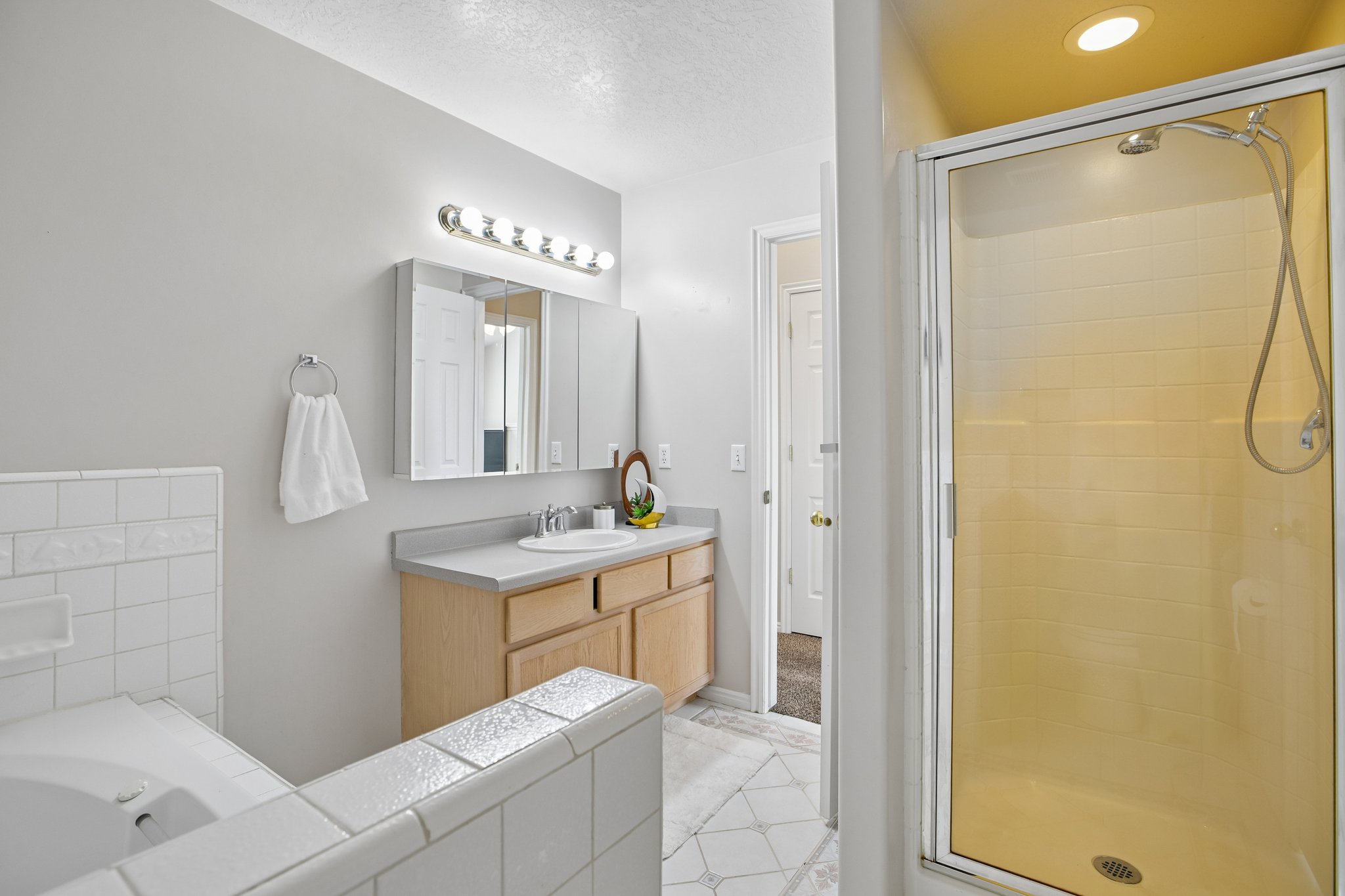 Bathroom featuring a glass-enclosed shower, a built-in soaking tub with white tile surround, a single vanity with a laminate countertop, and overhead vanity lighting