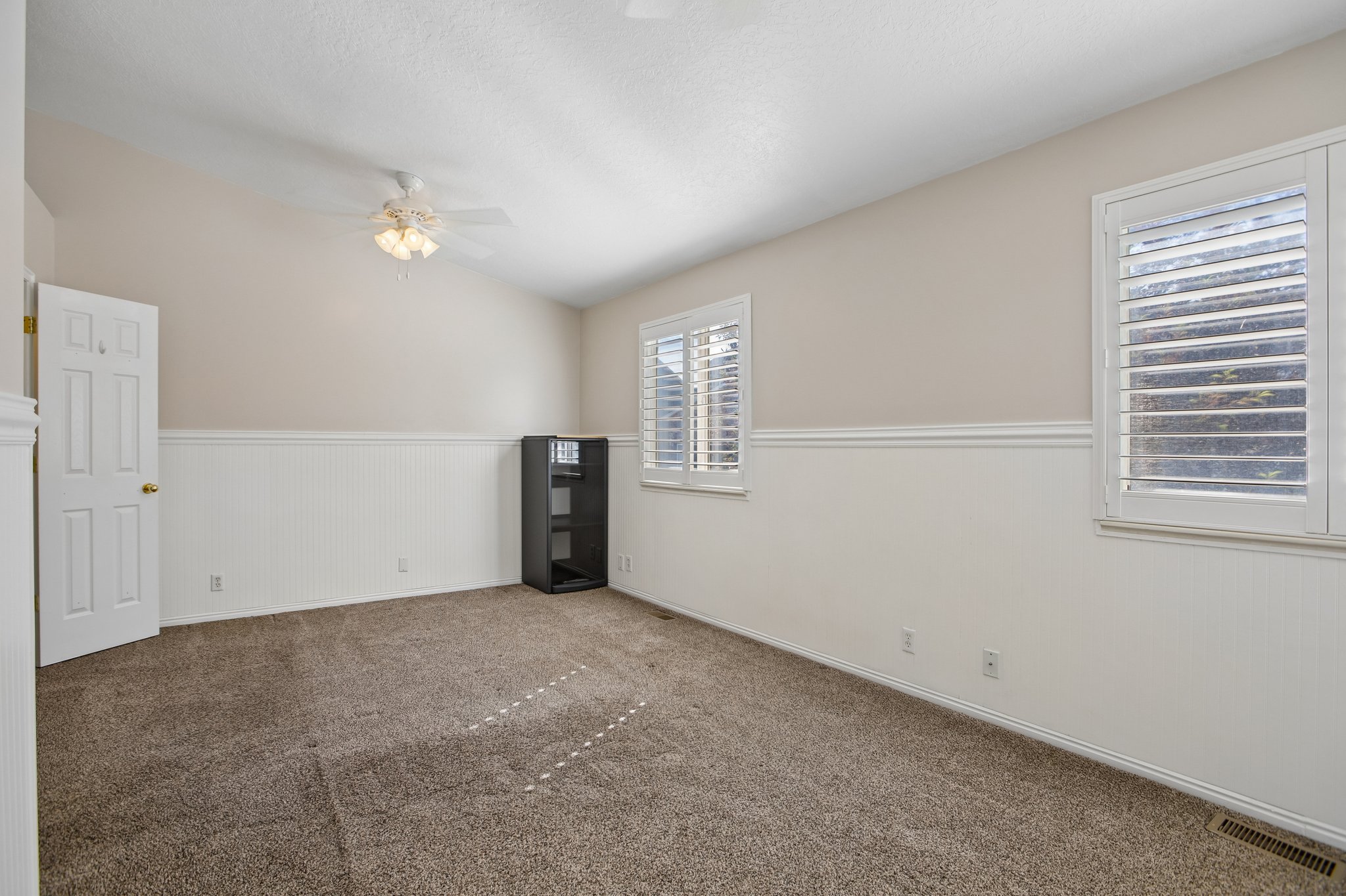 Carpeted room featuring a ceiling fan, two windows with plantation shutters, white wainscoting, and neutral-toned walls