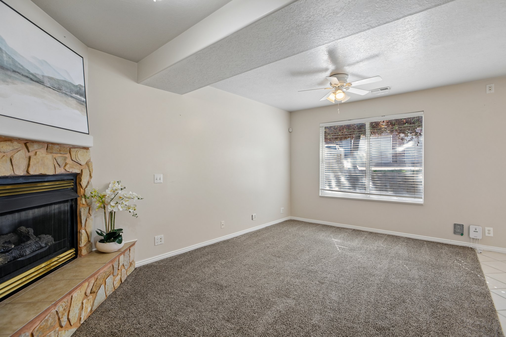 Spacious living area featuring a stone fireplace with a hearth, neutral carpeting, a ceiling fan with integrated lighting, and a large window with horizontal blinds