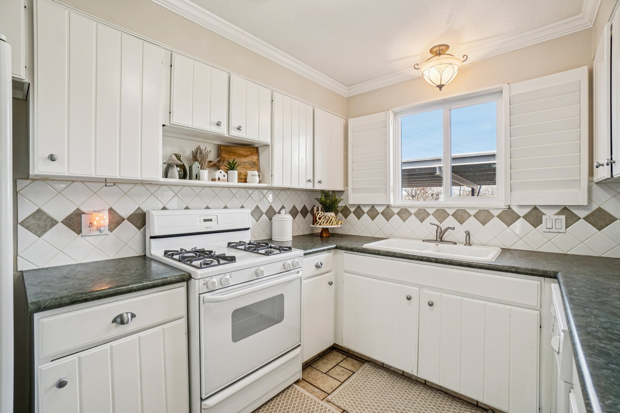 Kitchen featuring white paneled cabinetry, a diamond-patterned tile backsplash, dark stone countertops, a white gas range, and a white single-basin sink under a window with white shutters