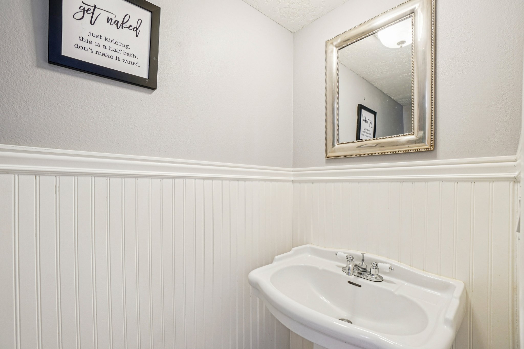 Pedestal sink with chrome faucet, complemented by white beadboard paneling and a decorative wall mirror