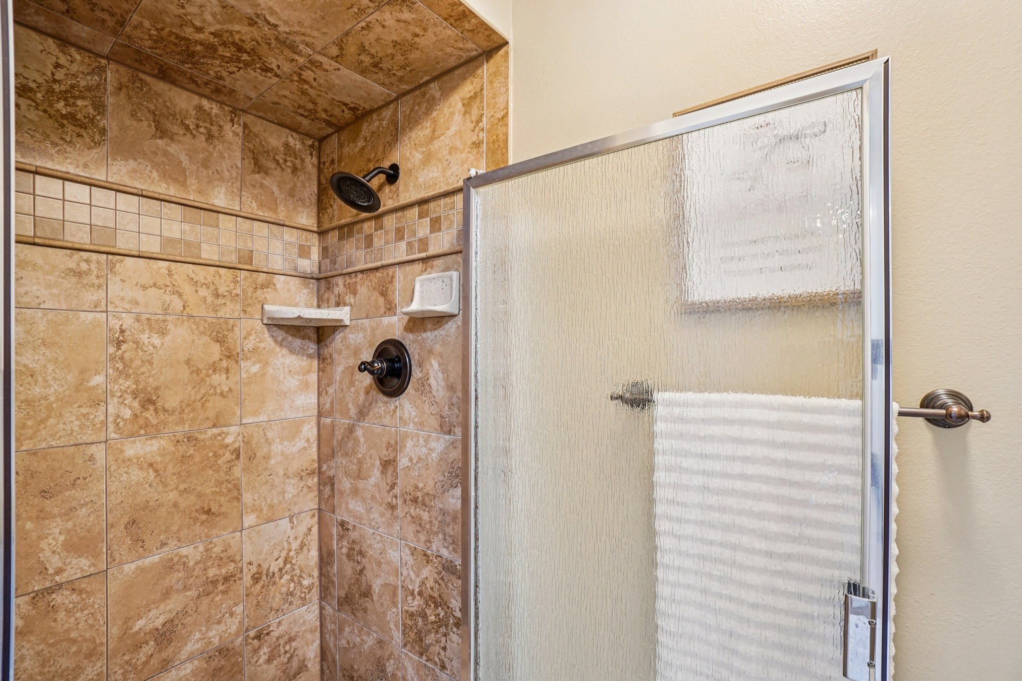 Tiled shower with a decorative mosaic band, bronze-finish fixtures, and a textured glass enclosure
